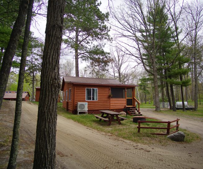 Cabin 6 front showning handrail steps access, air conditioning, picnic table in front, roads on both sides of cabin in a wooded setting